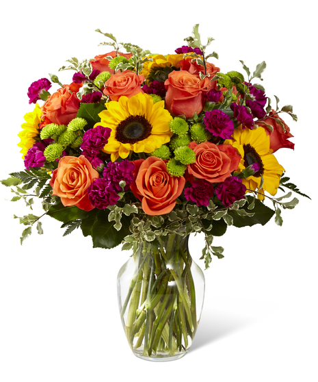The Color Craze bouquet features orange roses and summer sunflowers with lush foliage and seasonal accents.