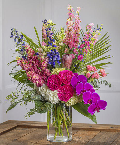 The Majesty Pastel Vase Arrangement features hydrangea, pink larkspur, protea, stock and exotic orchids in a tall, dramatic floral display